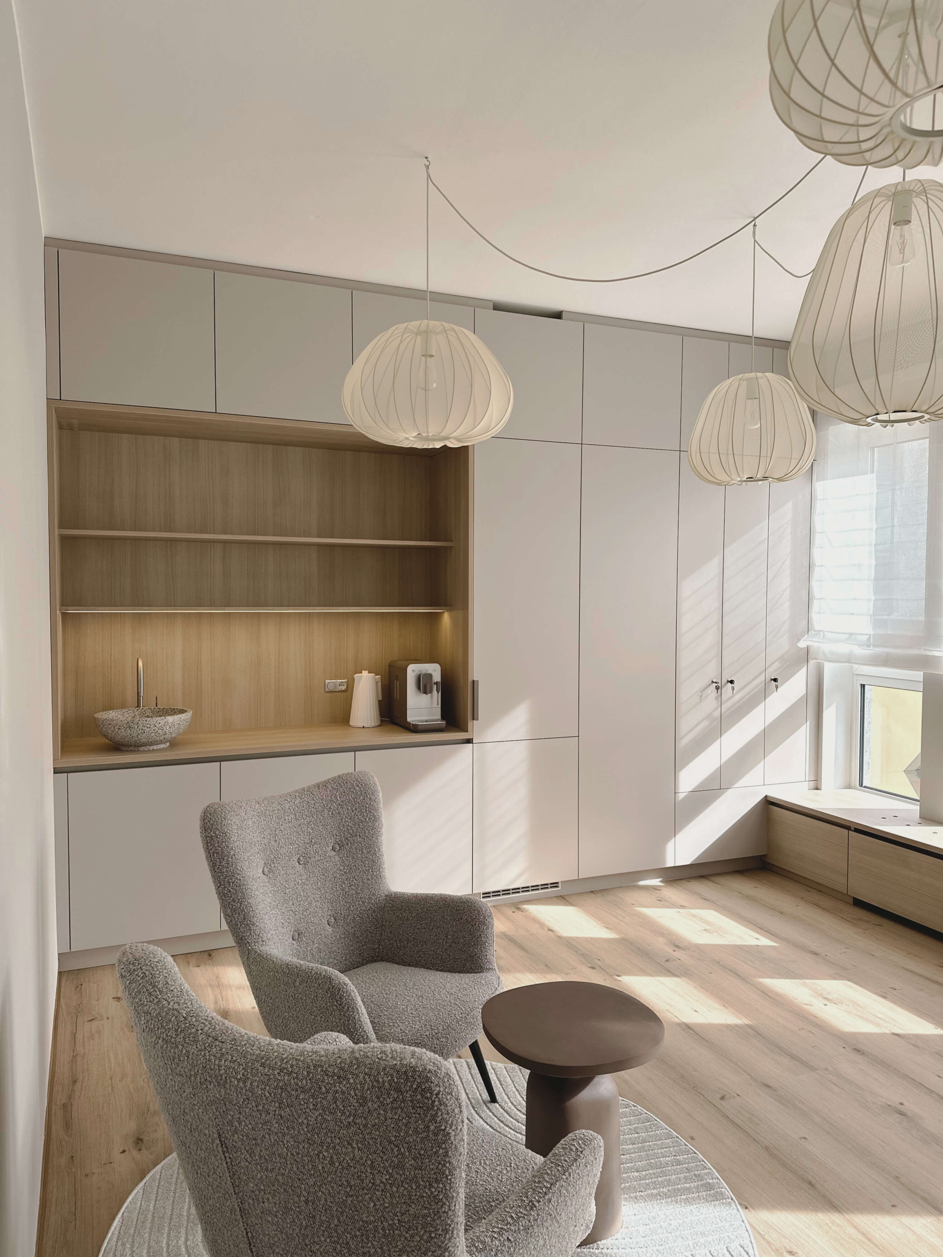 Foto einer modernen, lichtdurchfluteten Arztpraxis. Moderner Kastenverbau im Hintergrund in beige-grau. Moderne Textillampen und 2 komfortable helle Ohrensessel auf einem runden Teppich im Vordergrund. Eingerichtet im skandinavischen Stil, sehr wohnlich.